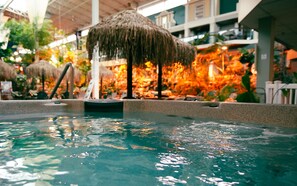 A pool area with clear water, a thatched umbrella, and indoor tropical plants.