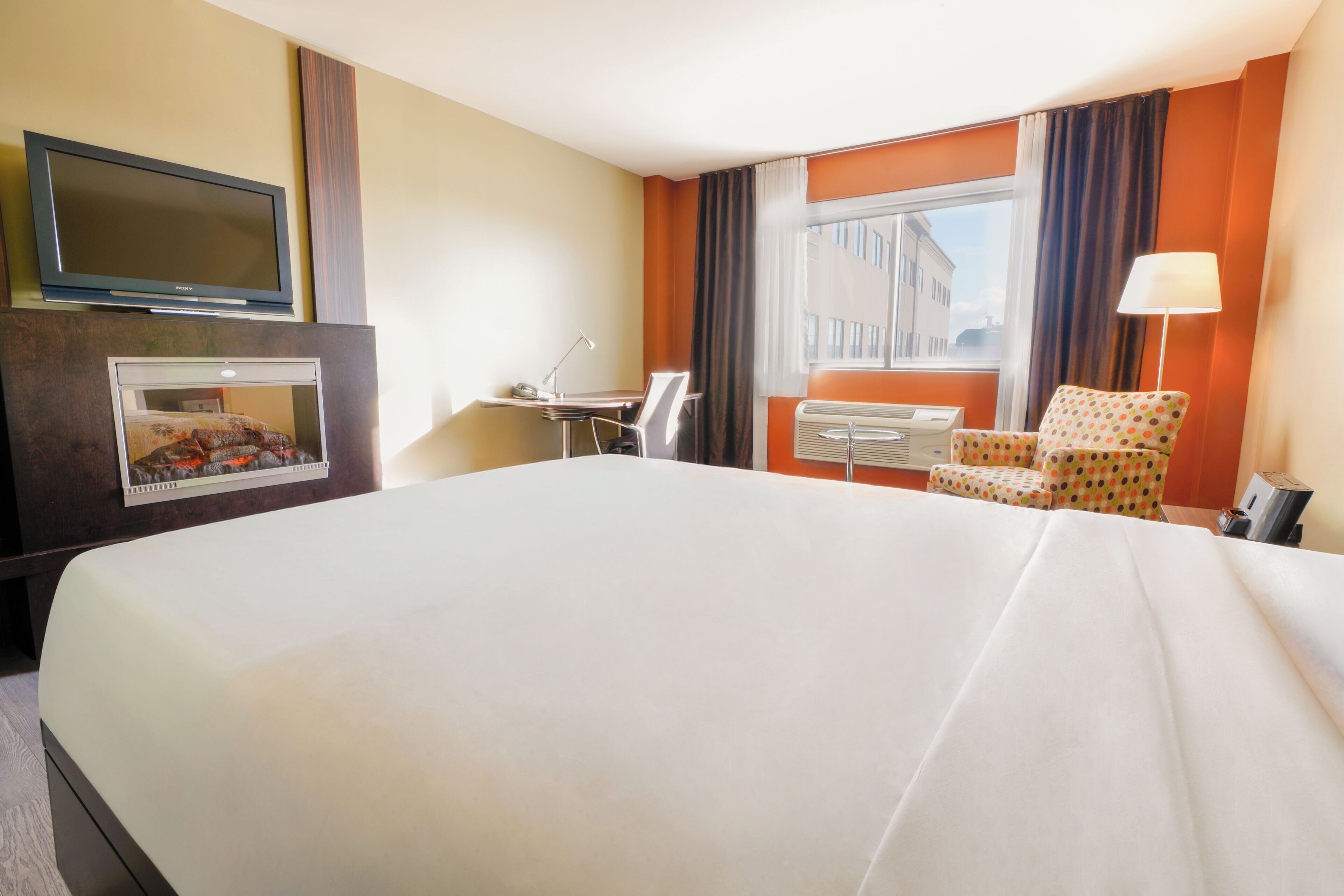 A hotel room with a large bed, a desk with a chair, a television, a window with curtains, and a lamp.