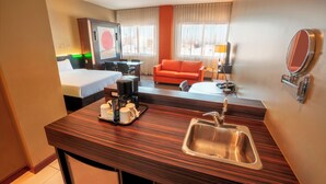 Habitación de hotel con cama, sofá, escritorio con lavabo, espejo y televisor.