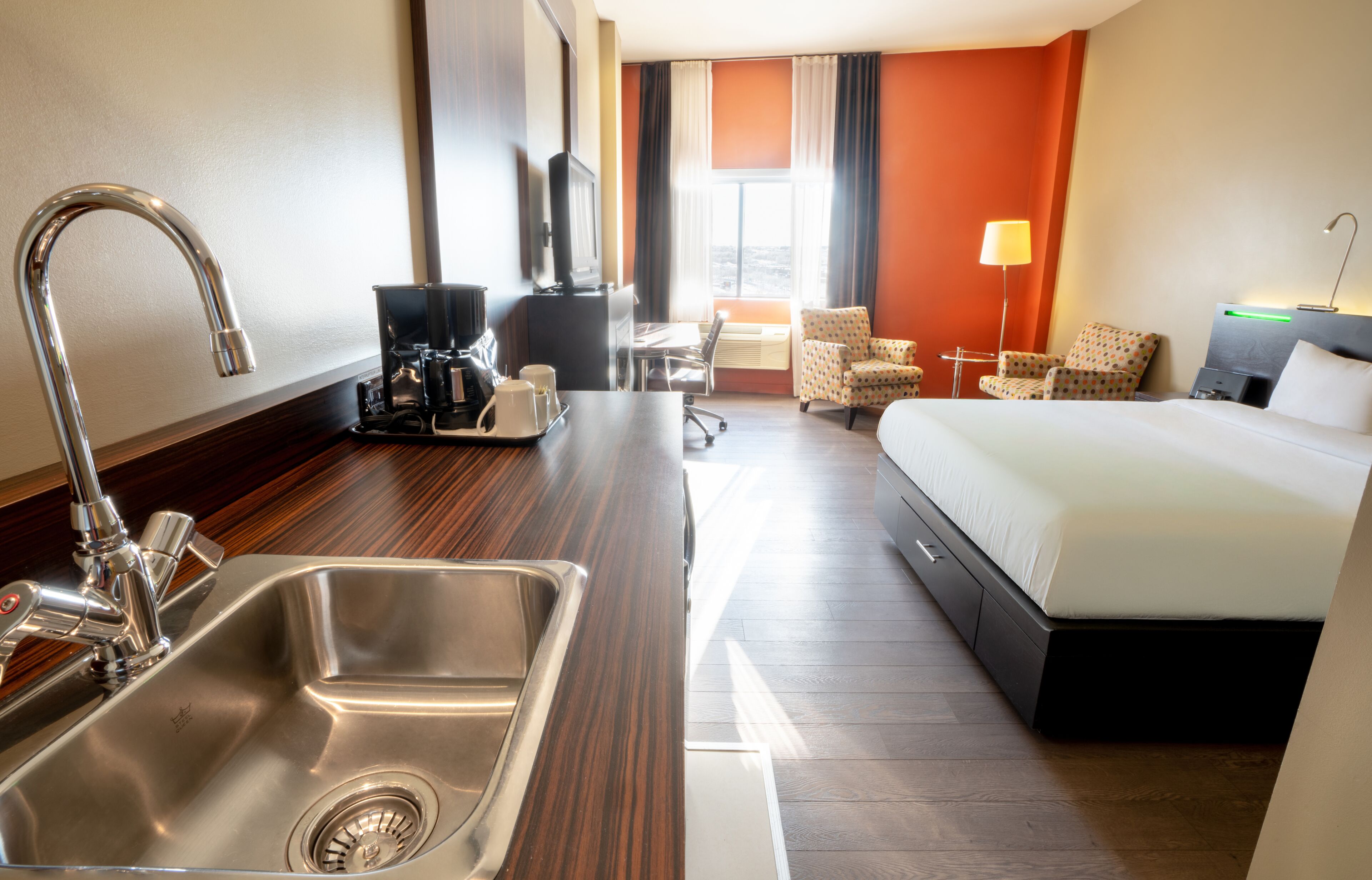 A hotel room with a bed, a desk with a coffee maker, a chair, a lamp, and a TV.