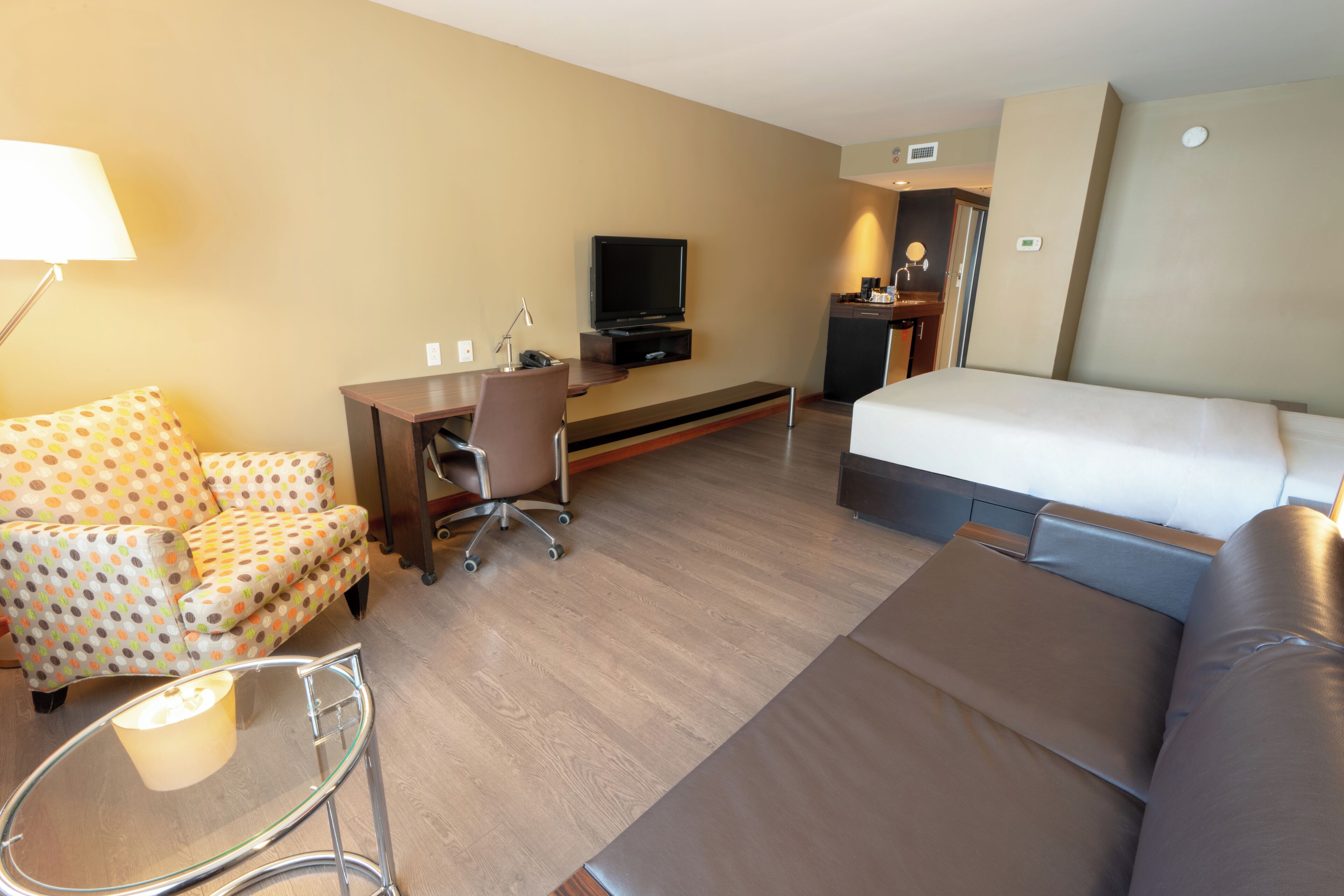 A hotel room with a bed, a sofa, a desk with a chair, a television, and a small kitchenette.