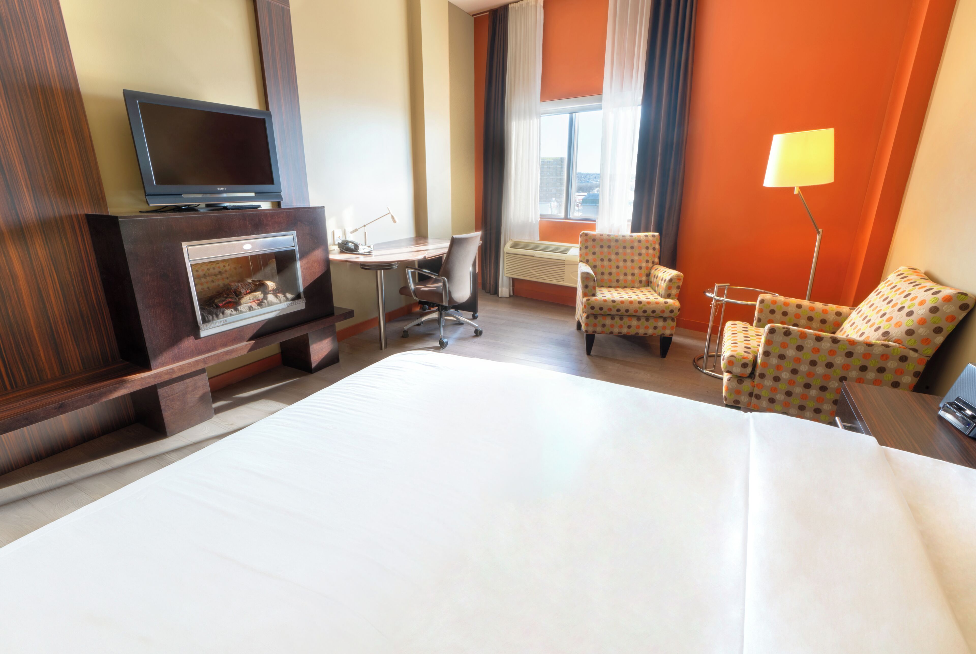 A hotel room with a bed, a desk with a chair, a television, and a fireplace.