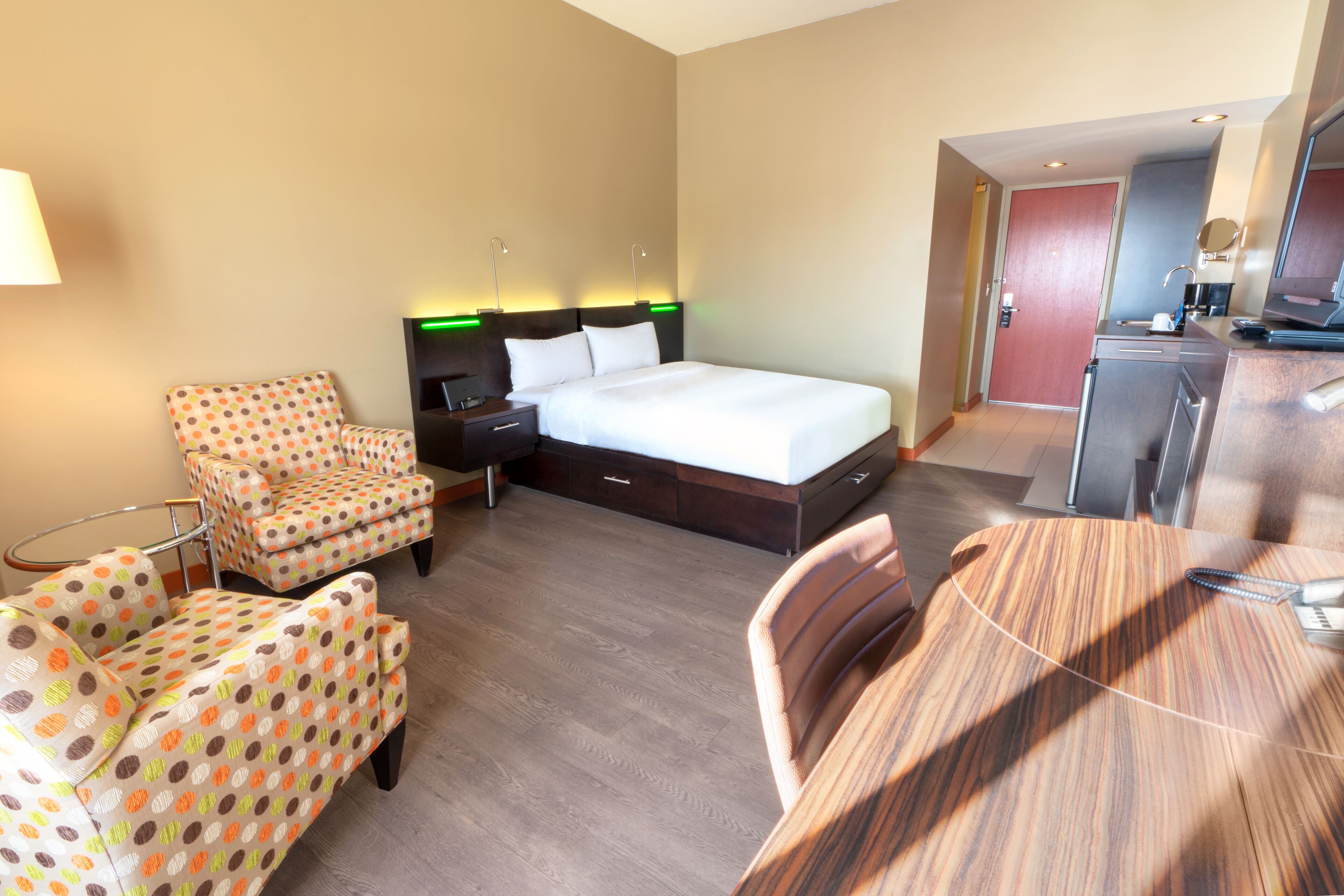 Quarto de hotel com cama, escrivaninha, cadeira, mesinha e cozinha compacta.