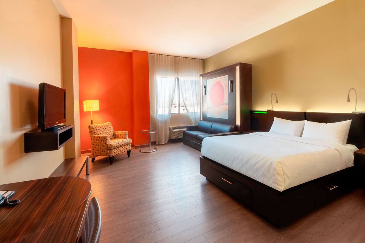 Habitación de hotel con cama, televisión, escritorio, silla y un ventanal grande con cortinas.