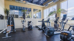 Un gimnasio con diverso equipo de ejercicio, ventanas amplias y un techo alto con conductos a la vista.