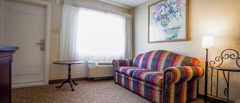 A hotel room with a patterned carpet, a colorful sofa, a small round table, a heater, and a framed floral artwork on the wall.