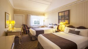 Quarto de hotel com duas camas, uma escrivaninha, uma cadeira, um abajur e um quadro na parede.