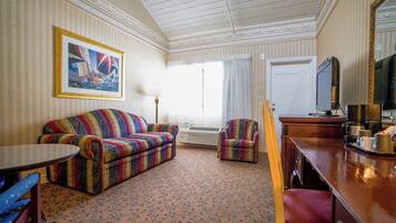 A hotel room with a striped wall, a sofa, a chair, a small table, a desk with a TV, and a large window with curtains.
