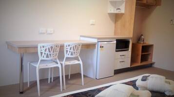 A compact kitchenette with a microwave, a small refrigerator, and a coffee maker on a shelf. There are two white chairs and a bed with rolled-up towels.