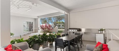 An open-plan living area with a dining table, chairs, and a kitchen area with a grill. There are potted plants and a sofa with red cushions.