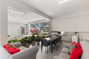 Espaço de estar integrado com mesa de jantar, cadeiras e área de cozinha com churrasqueira. Há plantas em vasos e um sofá com almofadas vermelhas.
