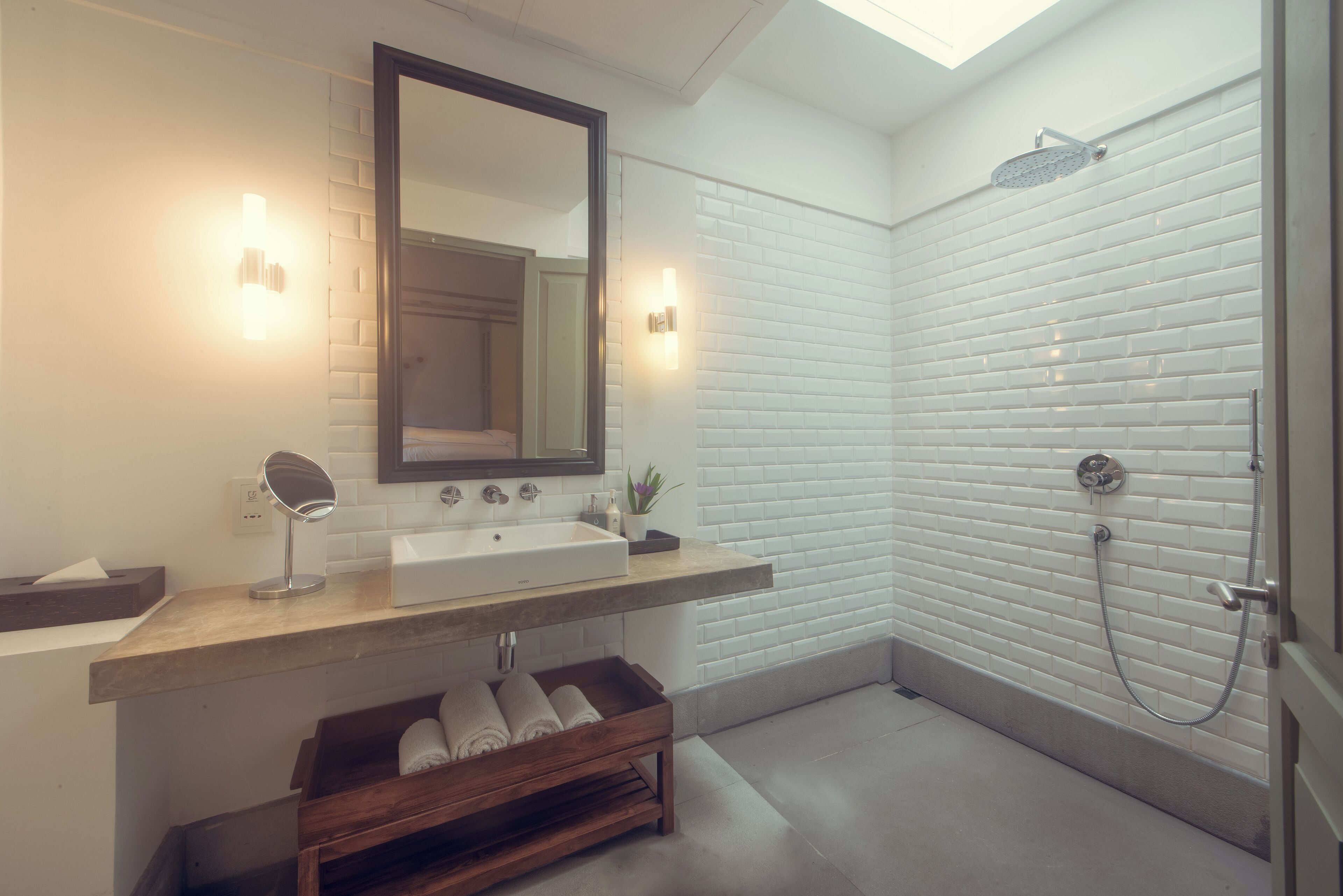 A modern bathroom with a white-tiled shower, a wooden bench with towels, a large mirror, and a countertop sink.