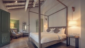 A four-poster bed with a wooden frame, a bedside table with a lamp, and a chair in a room with a green door and a mirror on the wall.