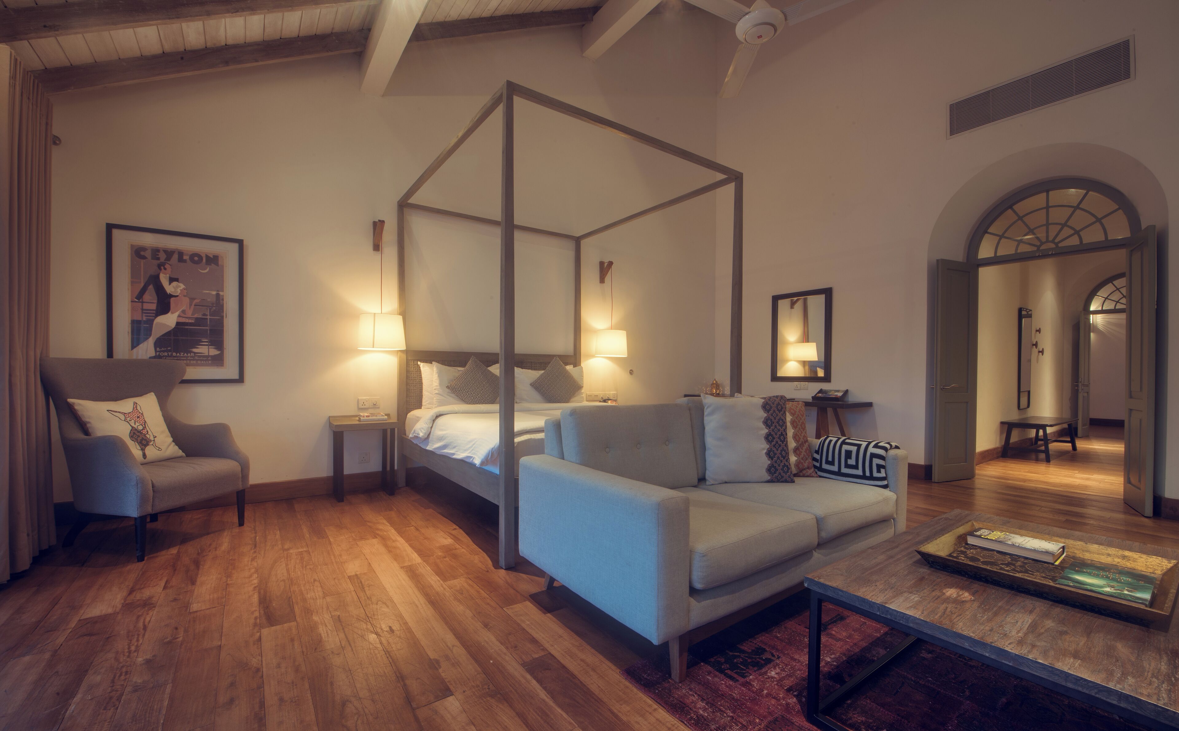 A four-poster canopy bed with a grey sofa and armchairs, wooden flooring, and a coffee table with a magazine.