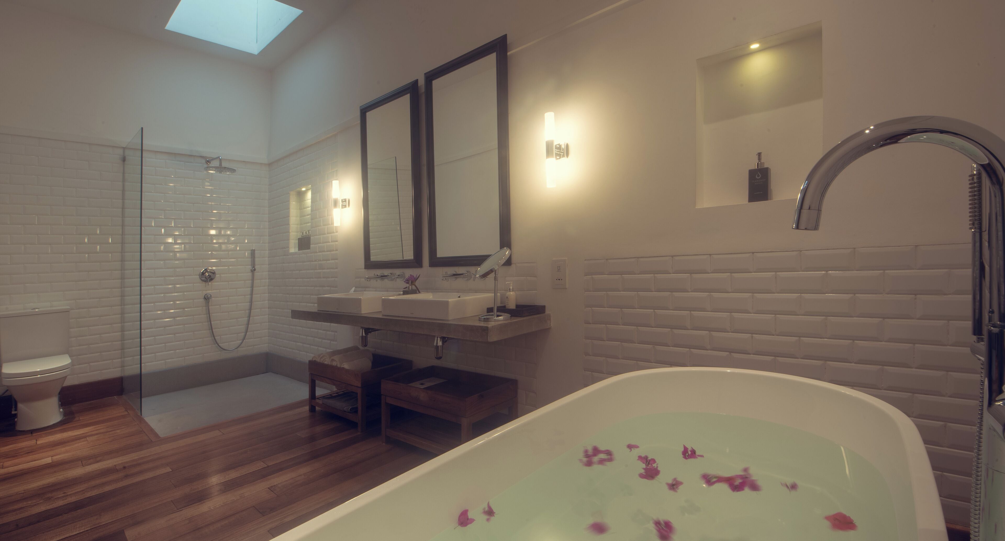 A modern bathroom with a glass-enclosed shower, a freestanding bathtub with rose petals, a double sink vanity, and a skylight.