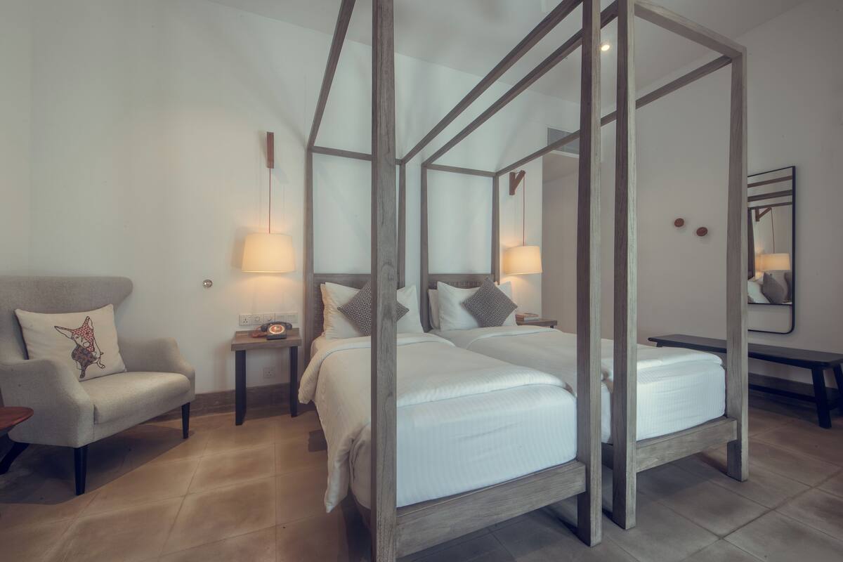 A four-poster bed with white linens, a wooden nightstand with a lamp, a chair with a cushion, and a mirror on the wall.