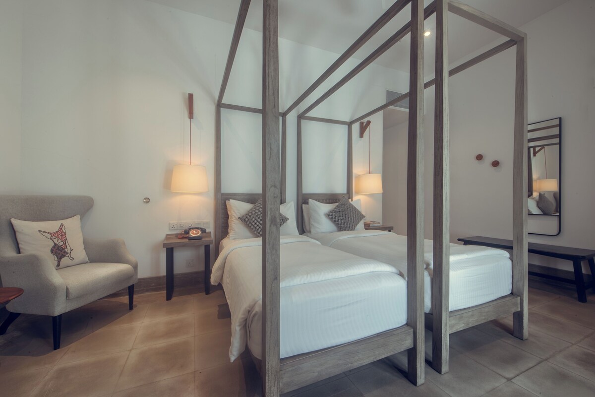 A four-poster bed with white linens, a wooden nightstand with a lamp, a chair with a cushion, and a mirror on the wall.