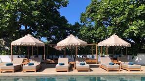 Un área junto a la piscina con sillones de descanso y sombrillas, rodeada de exuberante vegetación.