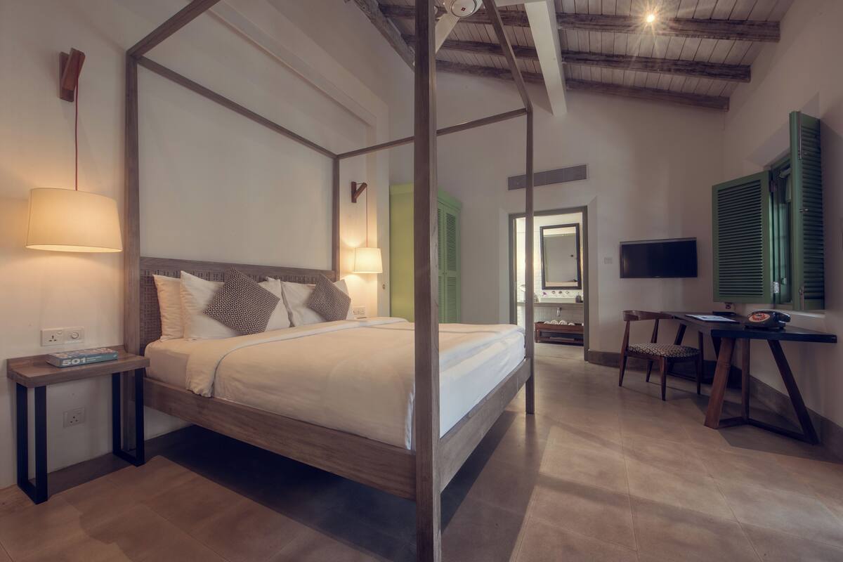 A four-poster bed with a wooden frame, a bedside table with a lamp, a desk with a chair, and a television on the wall.