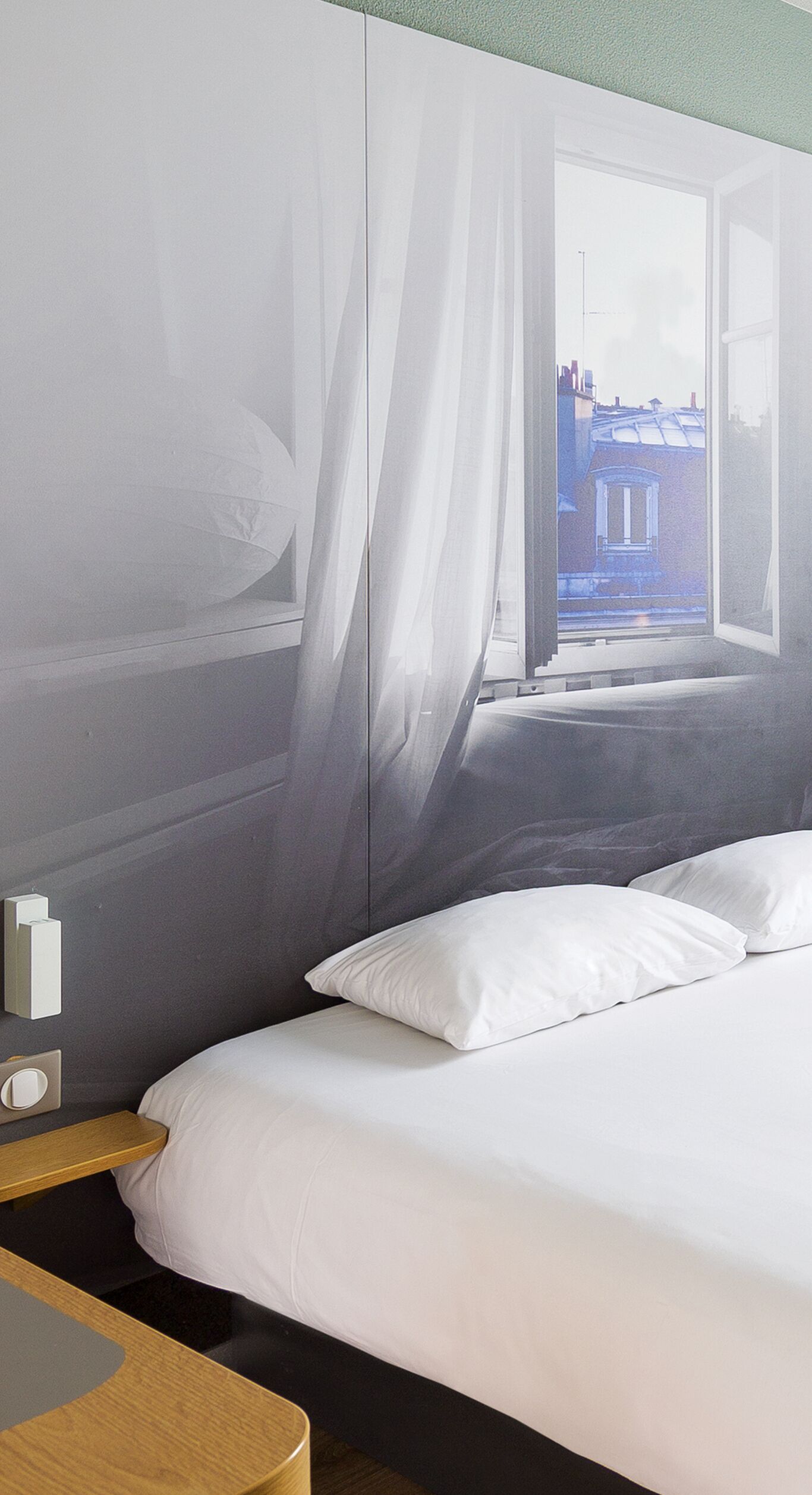 A modern hotel room with a large bed, a bedside table, and a window with sheer curtains.