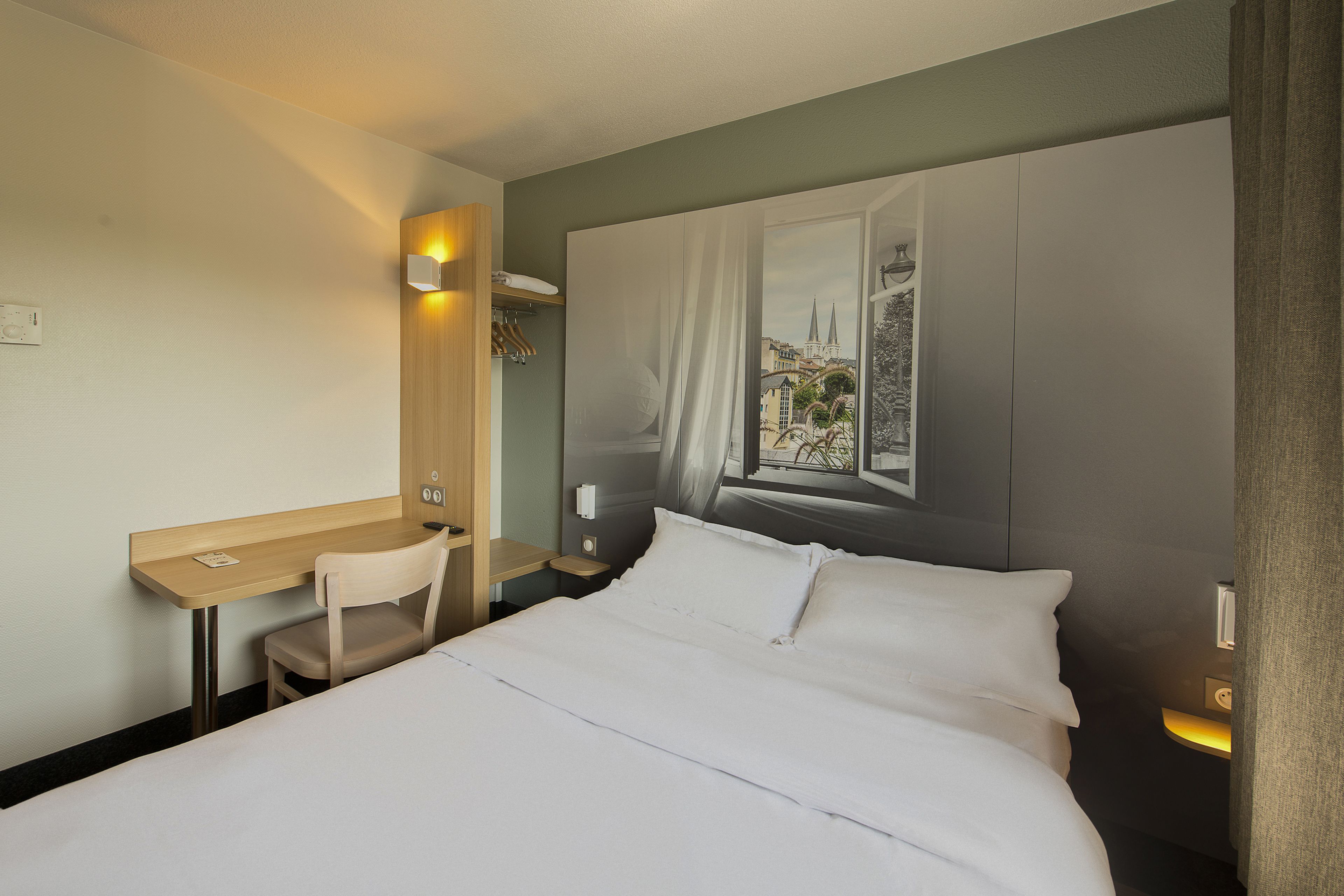 A hotel room with a bed, a desk, and a chair. There is a window with a view of buildings and a curtain.