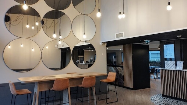 A modern interior with a long wooden table and four bar stools, surrounded by circular mirrors on the wall. - B&B HOTEL La Rochelle Centre les Minimes (La Rochelle)