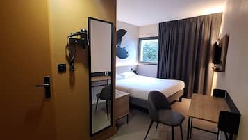 Une chambre d’hôtel compacte comprenant un lit, un bureau, une chaise, une petite table, un miroir, une fenêtre avec des rideaux et un téléviseur fixé au mur.