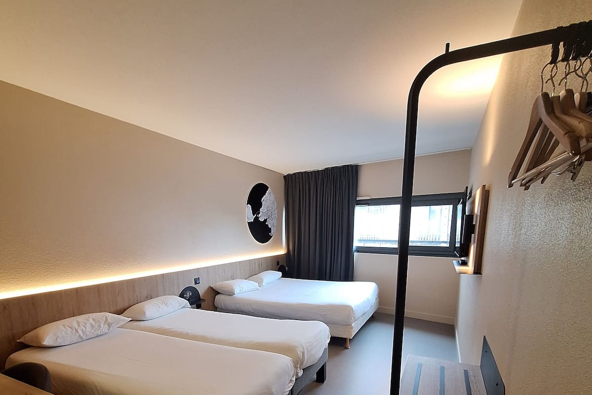 A compact hotel room with two single beds, a small desk, a chair, a wooden bench, and a wall-mounted TV.