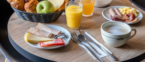 A breakfast table with croissants, bread, an apple, a glass of orange juice, scrambled eggs, bacon, cheese, and a cup of coffee.