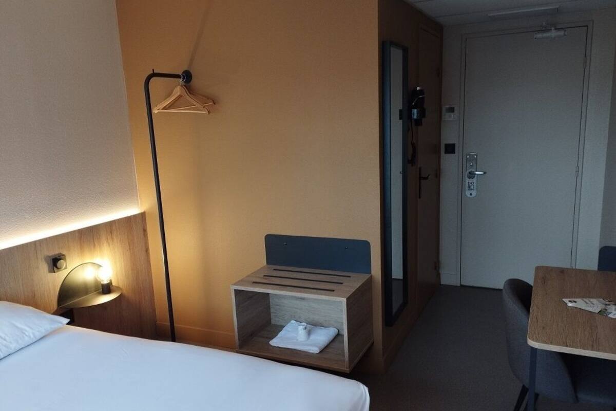 A hotel room with a bed, a bedside table with a lamp, a small wooden chest, a desk, and a chair.