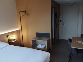Una habitación de hotel con una cama, una mesita de noche con una lámpara, un pequeño cofre de madera, un escritorio y una silla.