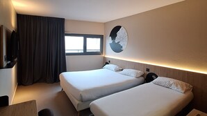 A hotel room with two beds, a desk, a chair, a TV, a window with curtains, and a wall-mounted fan. - B&B HOTEL La Rochelle Centre les Minimes (La Rochelle)