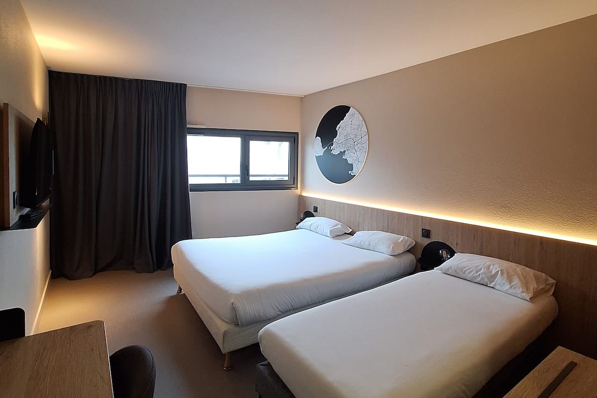 A hotel room with two beds, a desk, a chair, a TV, a window with curtains, and a wall-mounted fan.