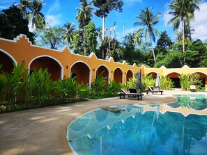 Zona de piscina en un resort, con piscina cristalina, sillas de descanso y un edificio amarillo con arcos.