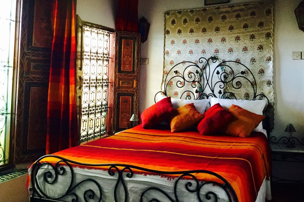 Cama com estrutura de metal e colcha vermelha e laranja.