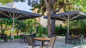 Un área de patio al aire libre con mesas y sillas bajo sombrillas, rodeada de follaje y árboles.