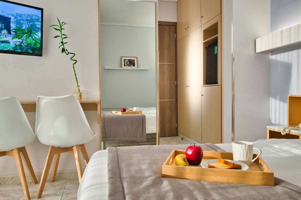 A hotel room with a bed, a desk with chairs, a small table with a picture, a plant, and a tray with fruit and a mug.
