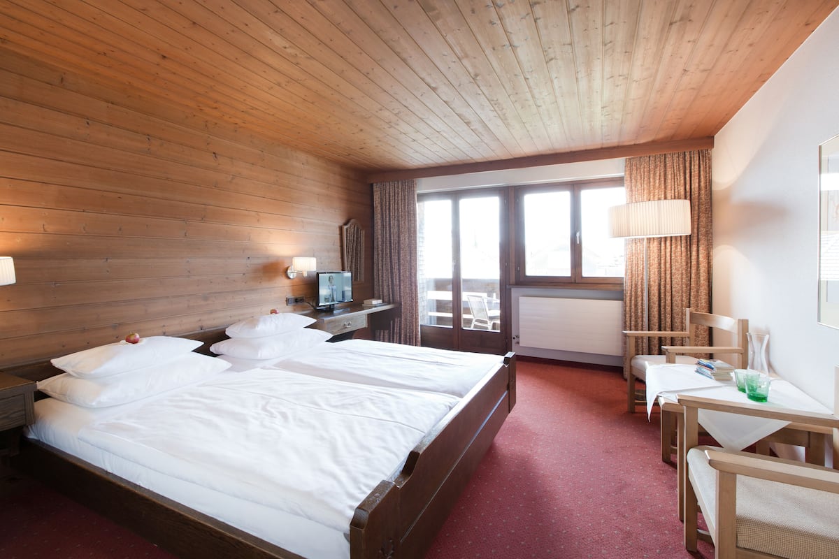 Habitación de hotel con techo de madera, una cama grande con ropa de cama blanca, un escritorio con lámpara y vista al exterior a través de una puerta corrediza.