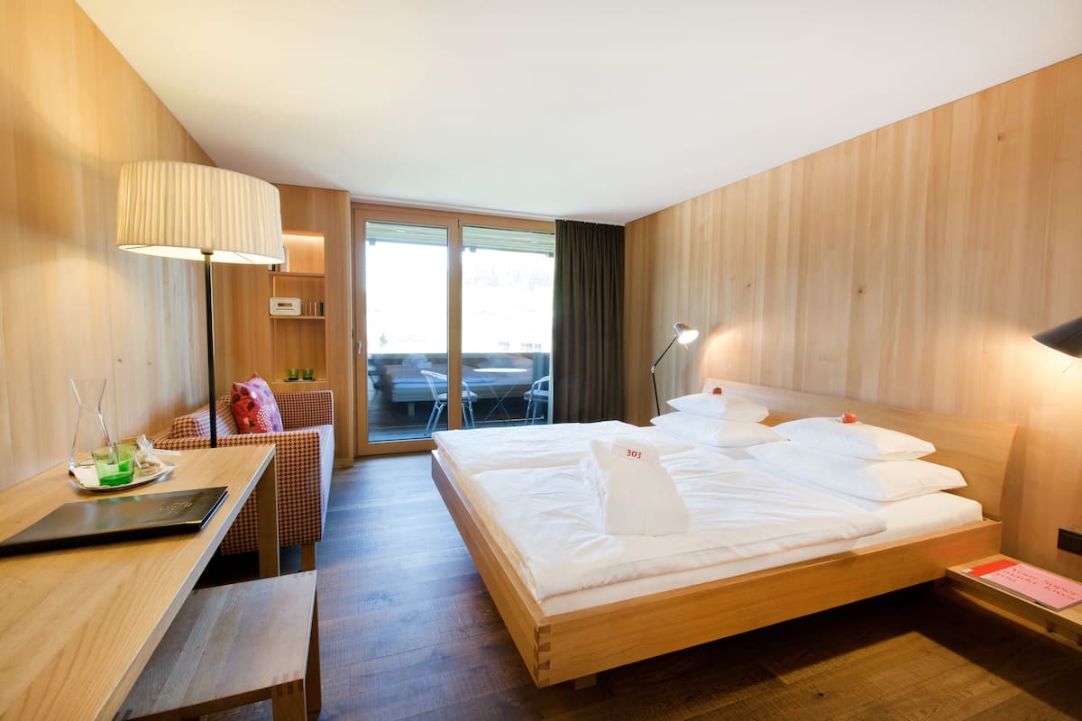 Habitación de hotel con una cama de madera, un escritorio con una computadora portátil, una silla y vistas al exterior a través de una puerta corredera.