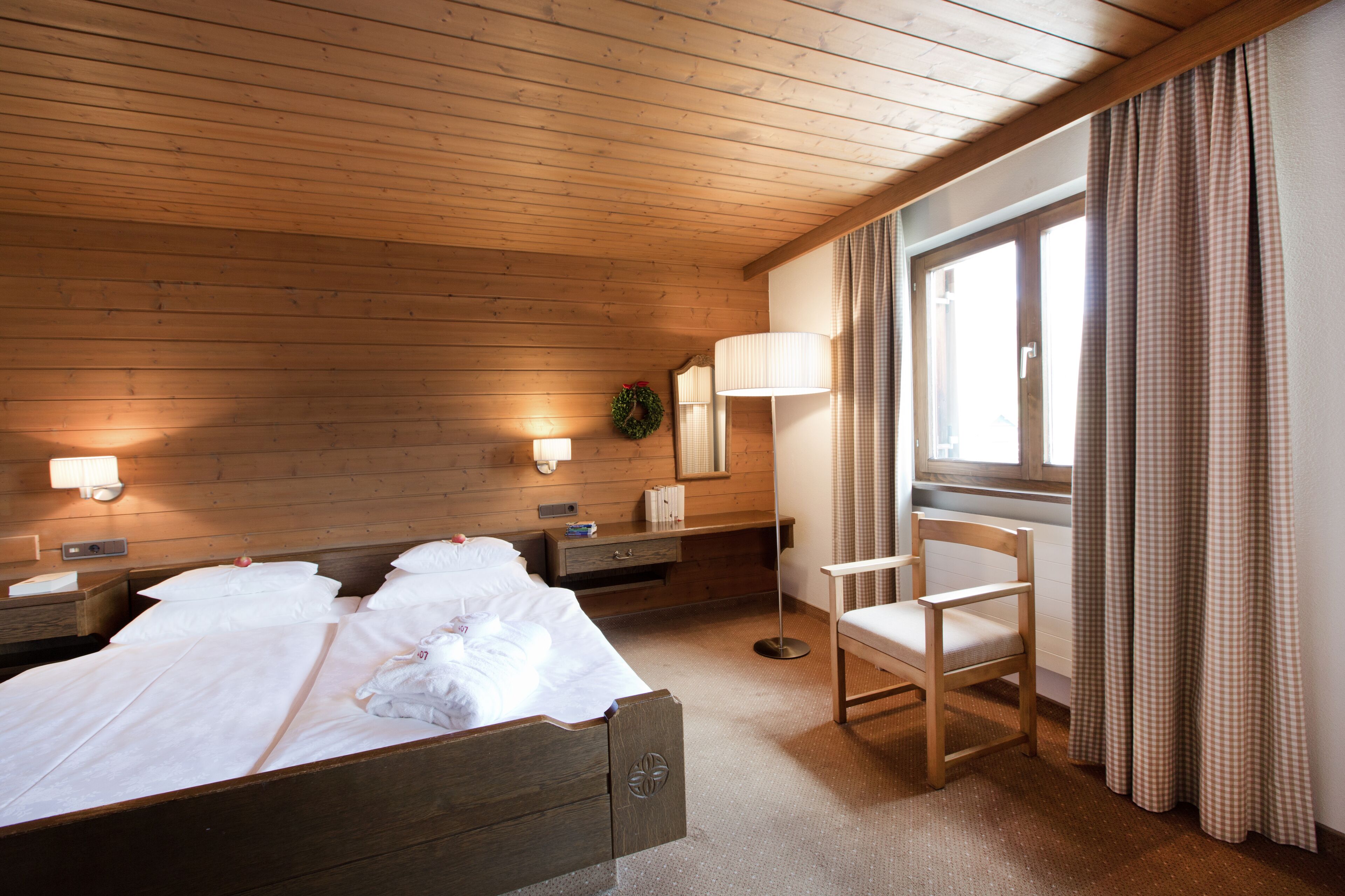 A hotel room with a wooden ceiling, a bed with white linens, a bedside table with a lamp, a chair, and a window with curtains.