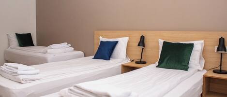 Dos camas individuales con sábanas blancas, una almohada azul, una almohada verde y una lámpara de escritorio negra.