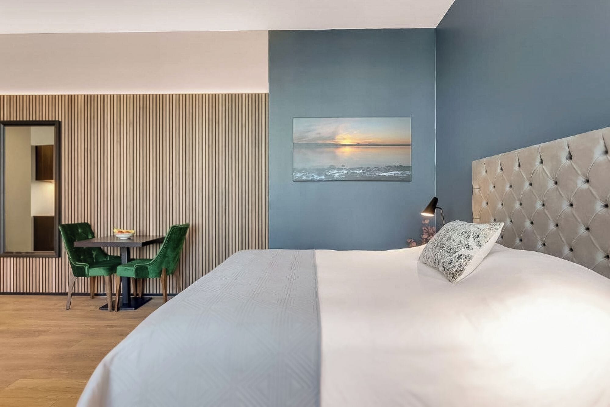 A hotel room with a large bed, a green chair, a small table, and a painting of a sunset.