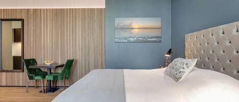 A hotel room with a large bed, a green chair, a small table, and a painting of a sunset.