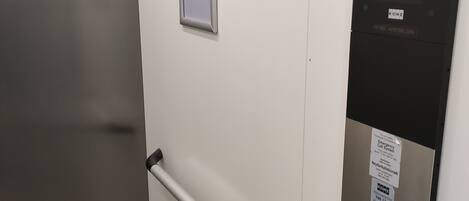 Panel de control de ascensor con instrucciones del sistema de llamada de emergencia, una puerta con manija y un ventanal pequeño.