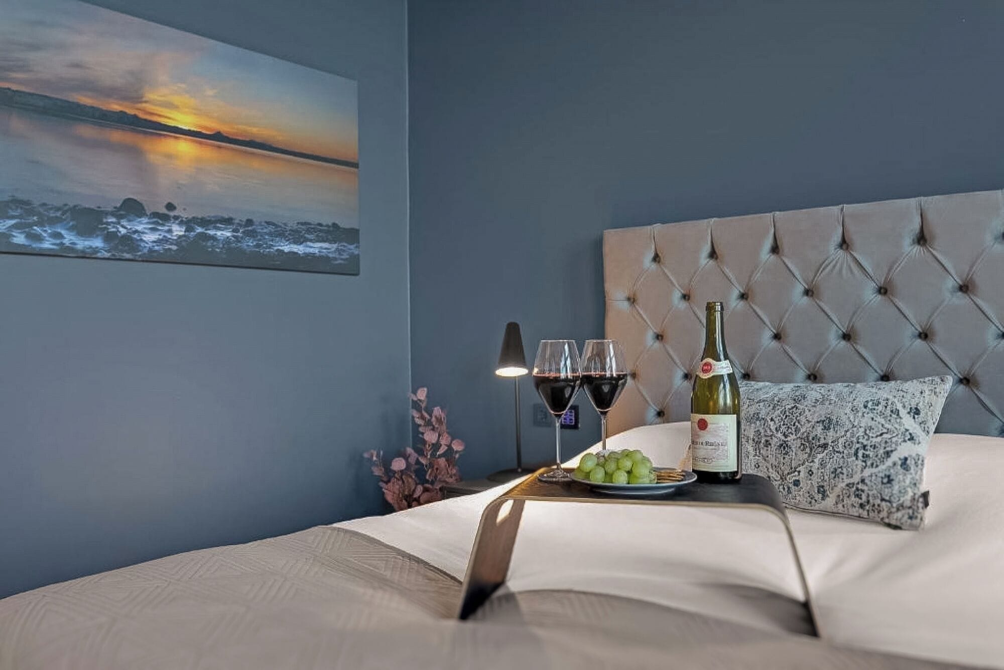 Un lit bien fait, avec une tête de lit, une bouteille de vin, deux verres et une assiette de raisins posée sur le lit.