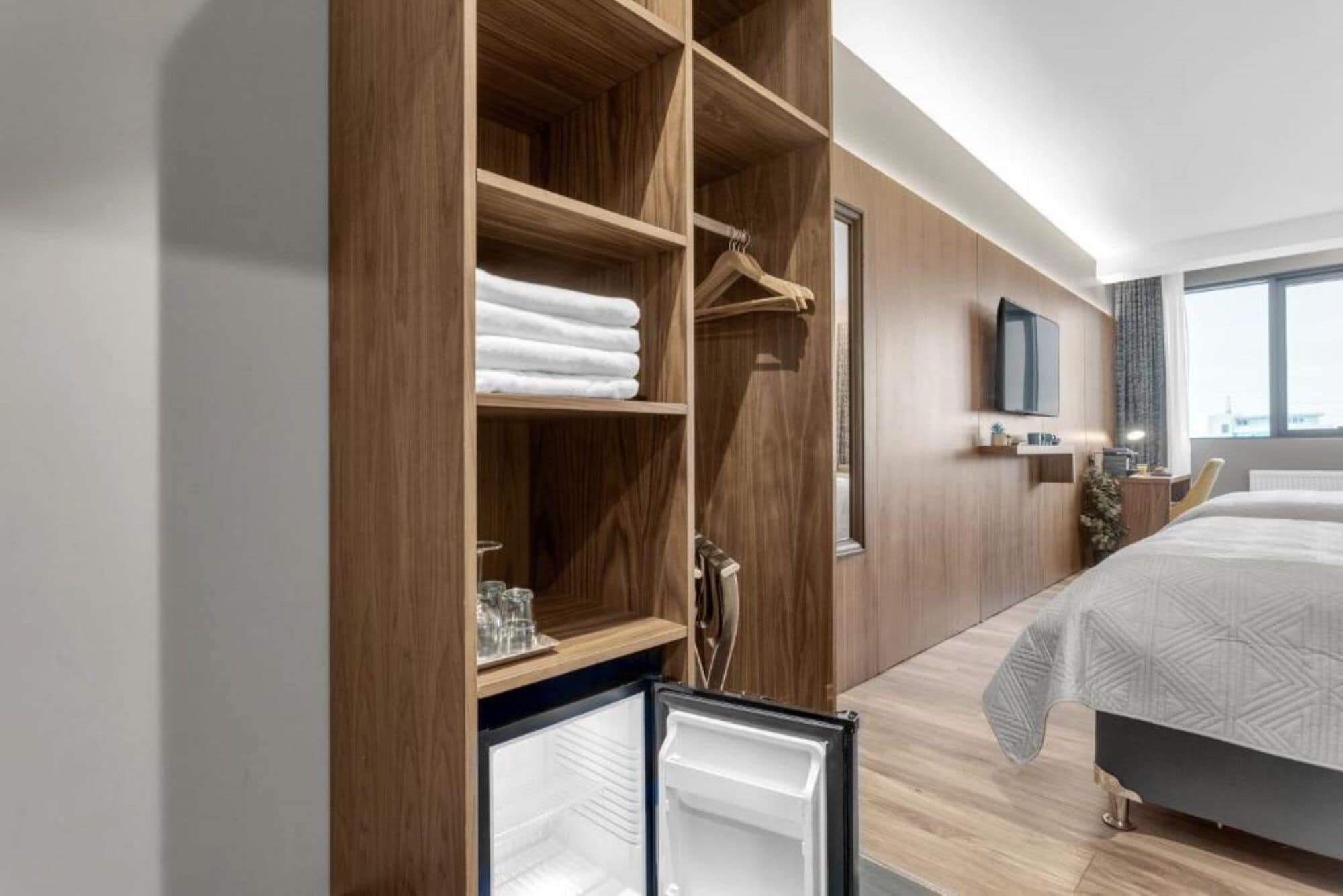 Et moderne hotelværelse med et træskab, en seng med et mønstret sengetæppe, et ophængt fjernsyn og et vindue med gardiner.