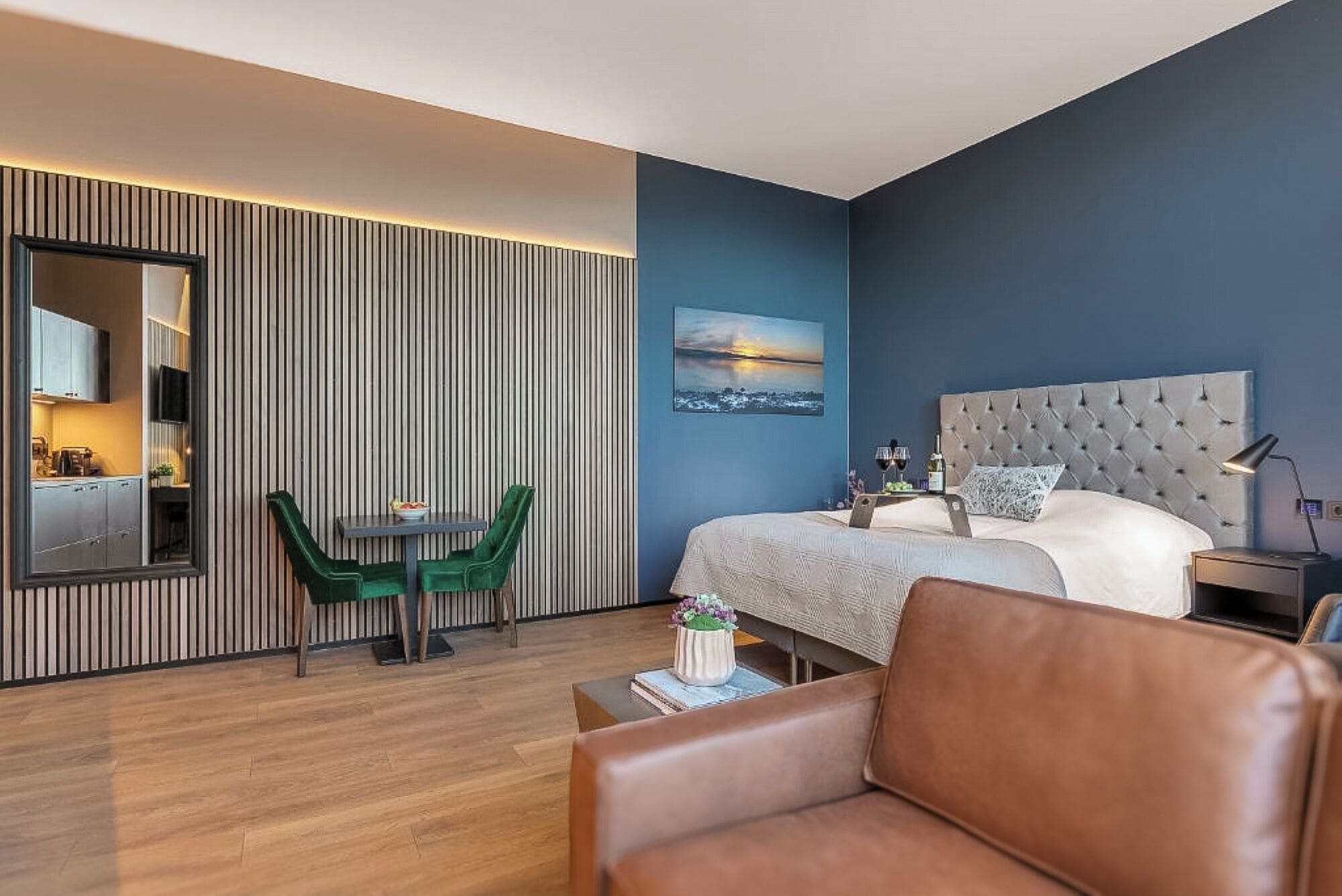 A modern hotel room with a bed, a dining table with chairs, a large mirror, and a cityscape painting on the wall.