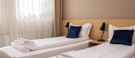 Dos camas individuales con sábanas blancas y almohadas azules, un cabecero de madera, una mesita de noche con lámpara y cortinas de gasa.