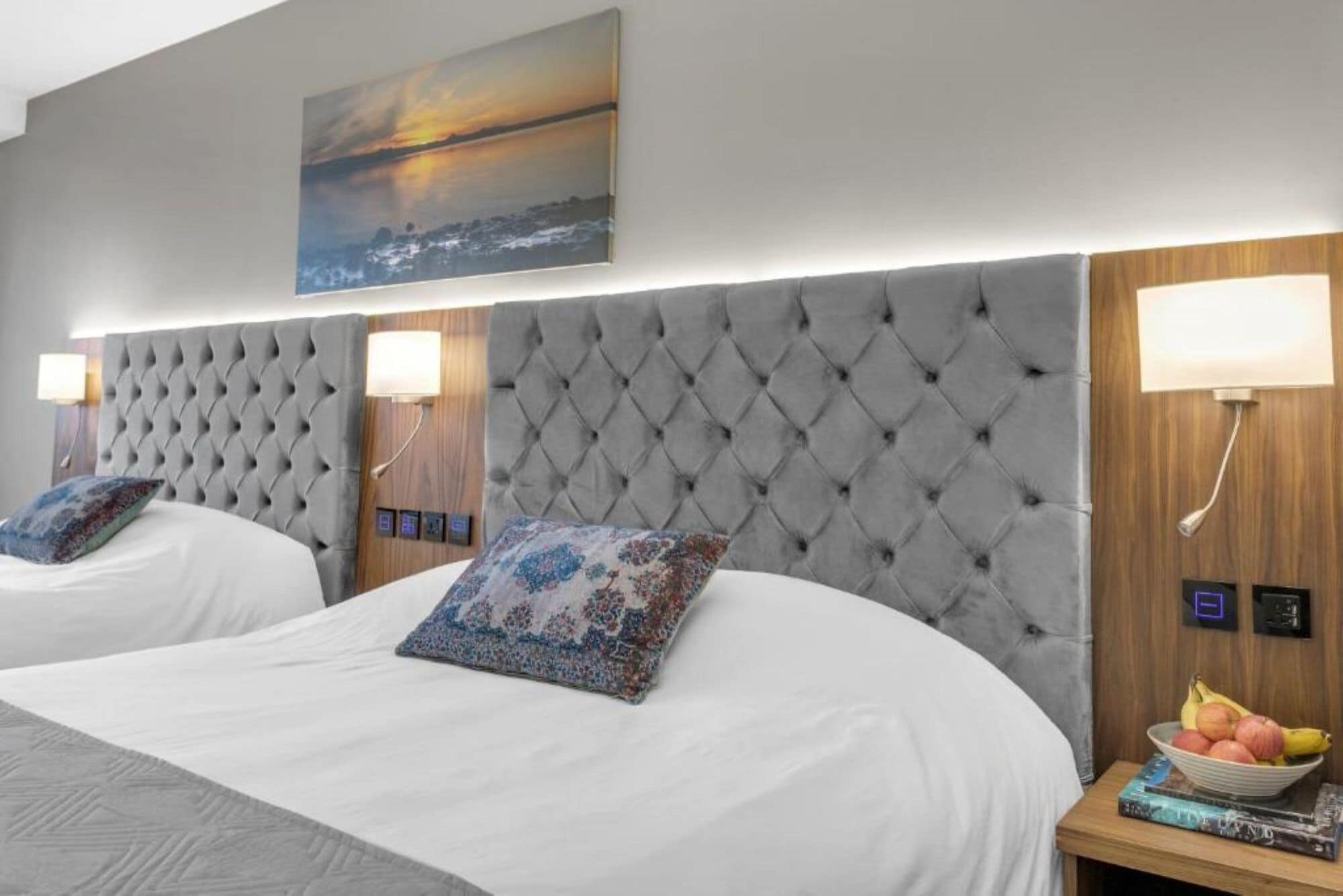 Habitación de hotel con dos camas, cada una con cabecero capitonado y una mesita de noche con lámpara y frutero.