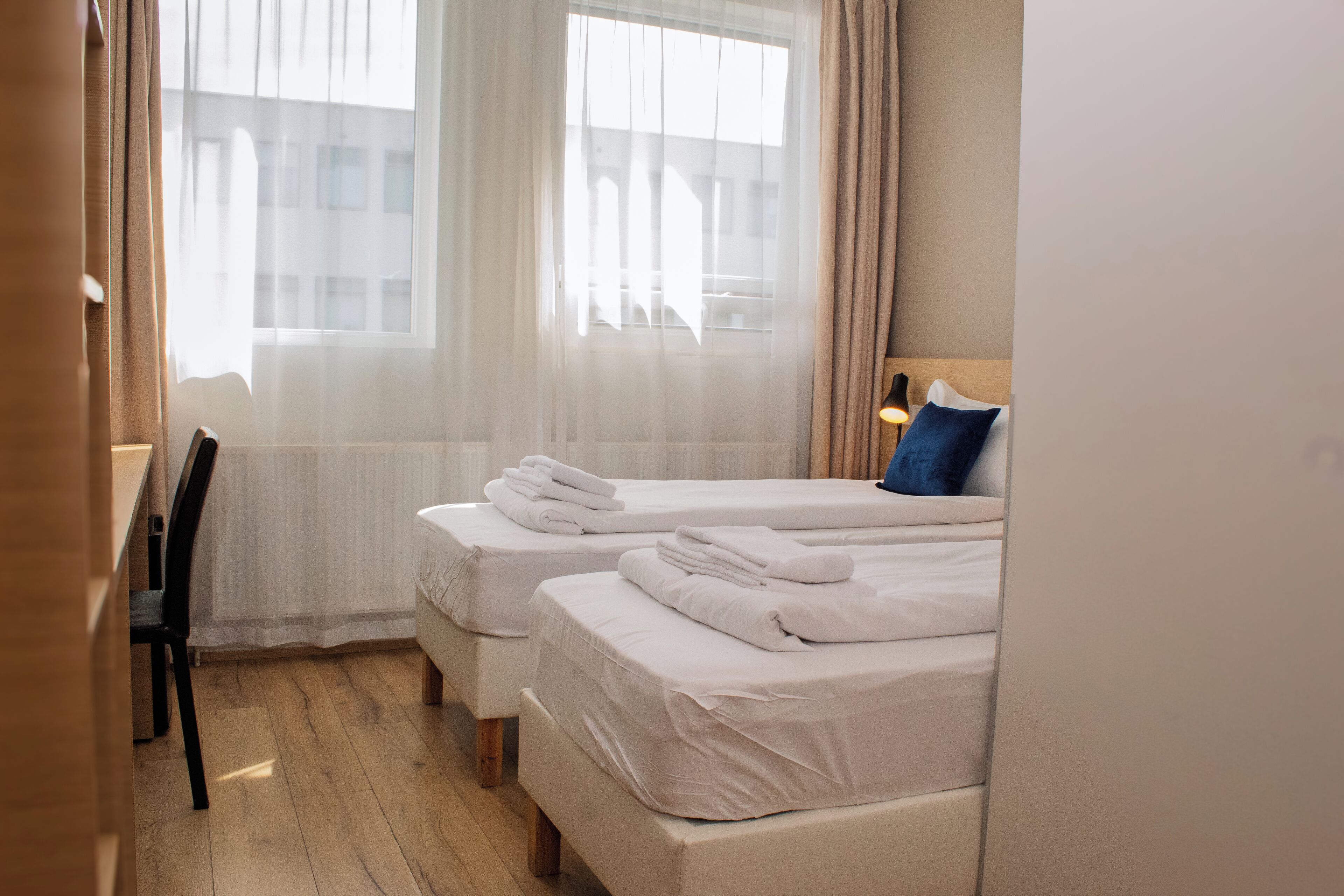 Une chambre d’hôtel avec deux lits, une chaise et une fenêtre avec des rideaux légers.